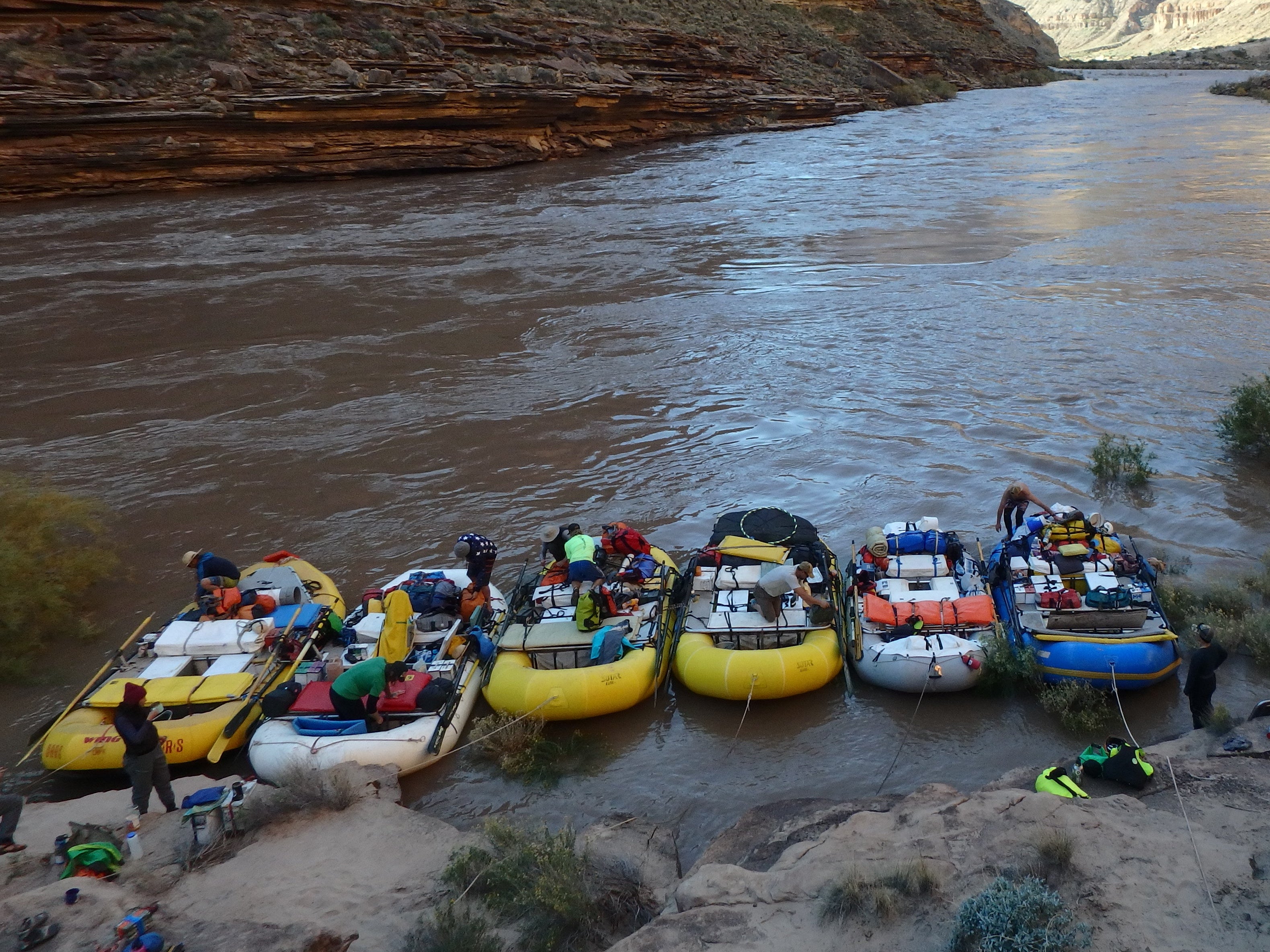 A scenic canyon river scene with several inflatable rafts and people on the sandy shore, with towering red rock canyon walls rising behind the water under a bright sky.